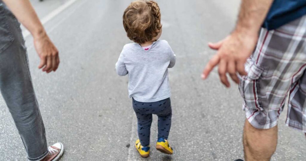 Toddler walking on city street with two adults nearby